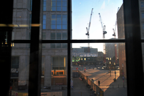 a view out of Alexanderplatz train station towards Alexanderplatz, with streaks of morning sun coming through commercial buildings and three cranes in the background