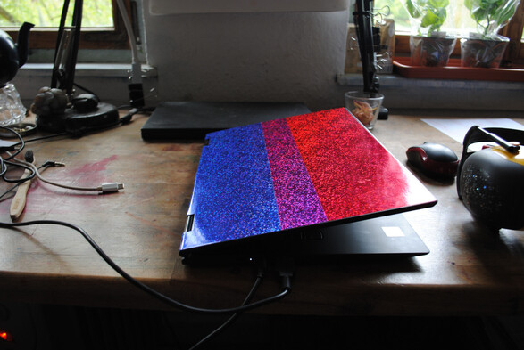 a half opened Thinkpad decorated with a strip each of blue, purple and red glitter foil, on a slightly cluttered wooden desk with another un-decorated Thinkpad and some plants in the background